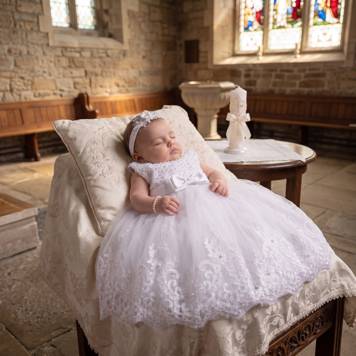 Bébé endormi dans une église portant une robe de cérémonie élégante avec haut perlé, ceinture à nœud et jupe en tulle brodée de dentelle.