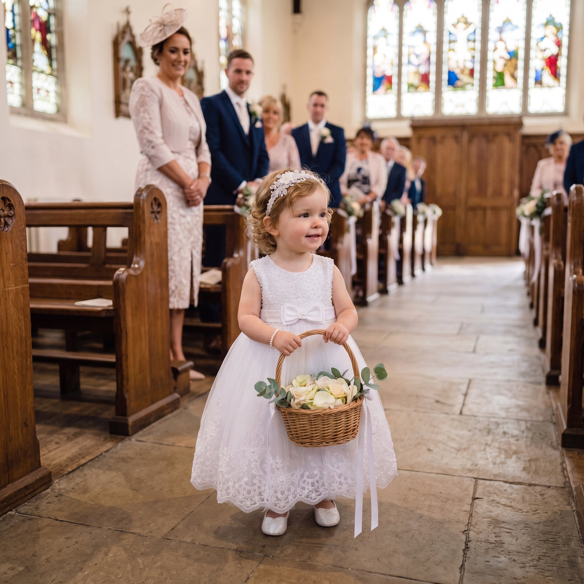 eune enfant tenant un panier de fleurs dans une allée d'église, vêtue d'une robe de cortège en tulle avec finitions en dentelle et détails de perles.
