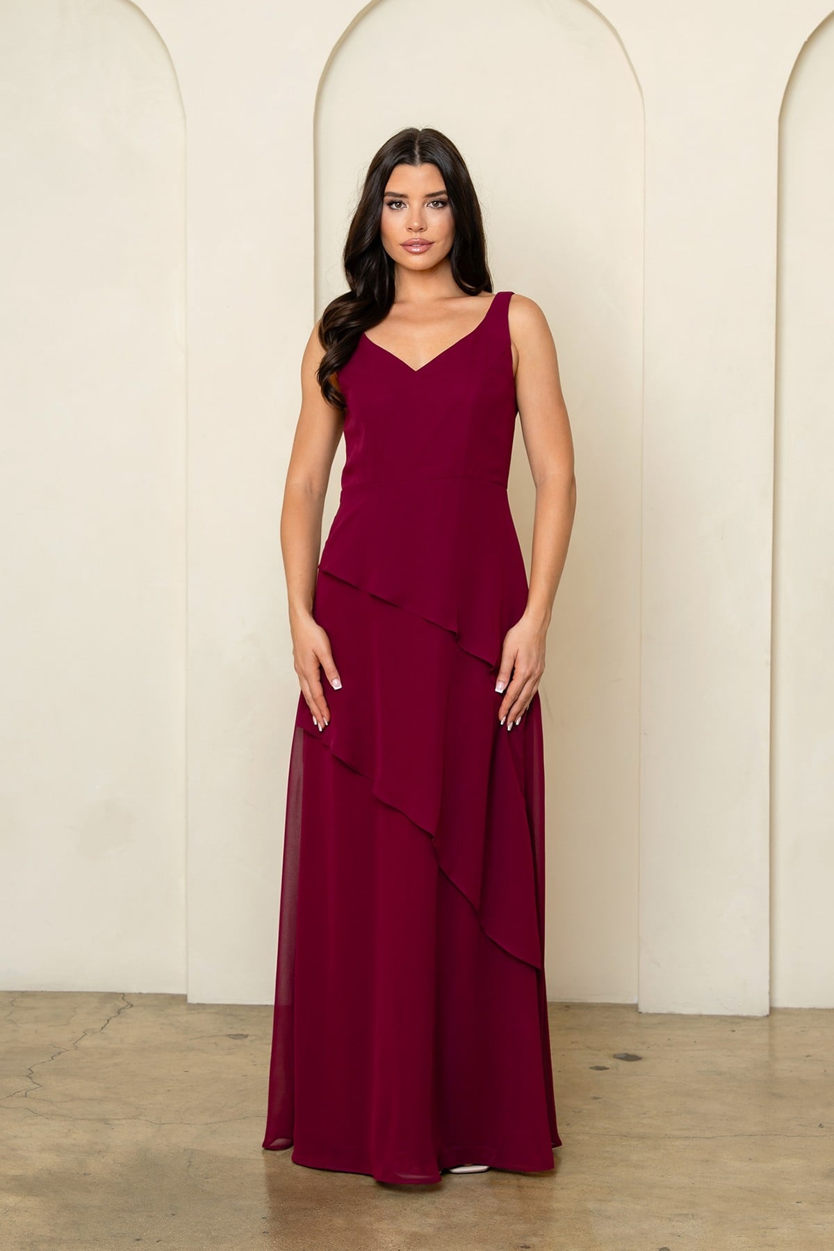 Robe de soirée longue bordeaux en mousseline fluide avec superposition style cape sur les épaules et jupe à volants asymétriques. Coupe élégante et classique pour occasions formelles.