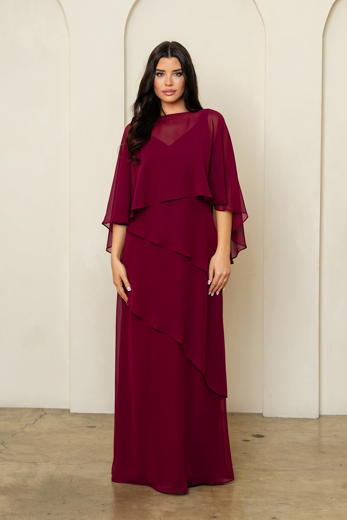 Robe de soirée longue bordeaux en mousseline fluide avec superposition style cape sur les épaules et jupe à volants asymétriques. Coupe élégante et classique pour occasions formelles.
