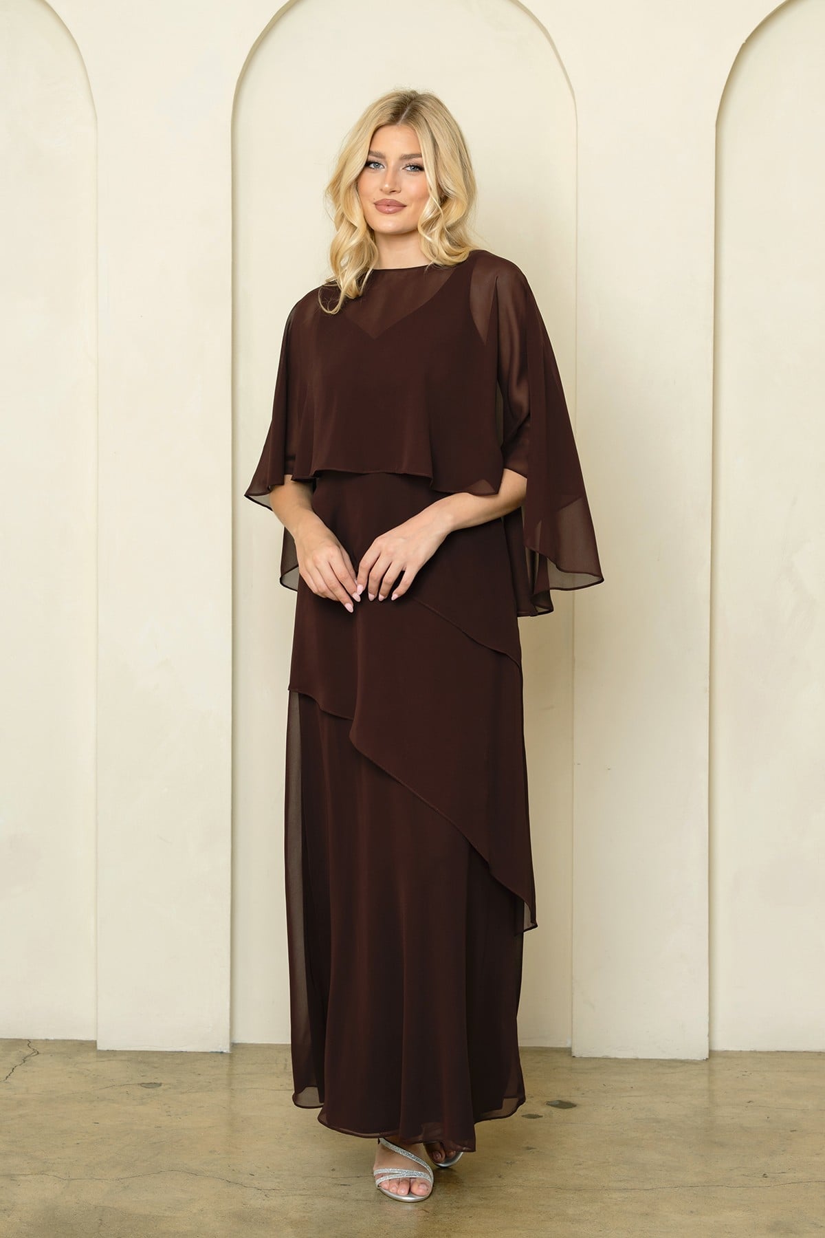 Robe de soirée longue brun en mousseline fluide avec superposition style cape sur les épaules et jupe à volants asymétriques. Coupe élégante et classique pour occasions formelles.