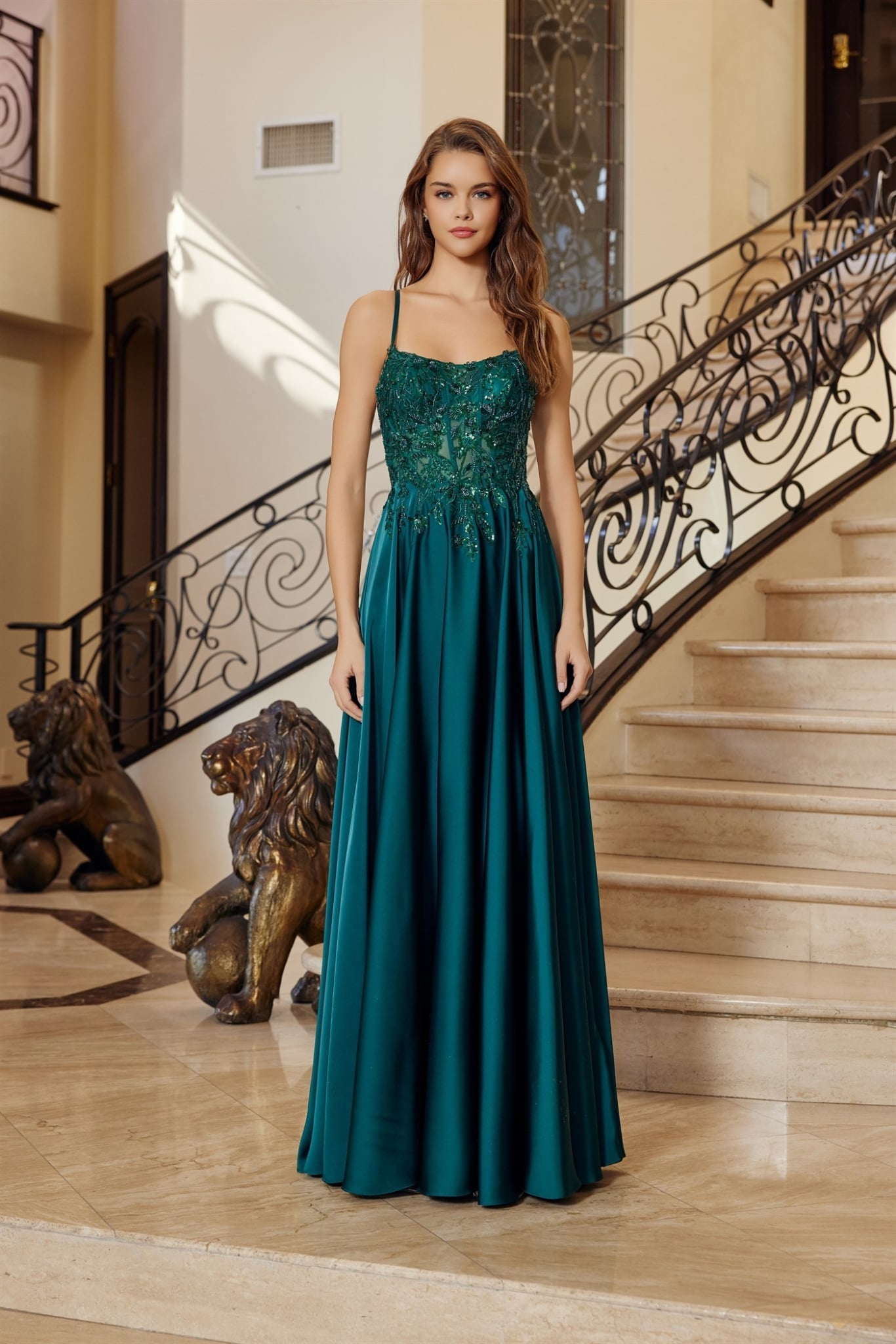 Robe de soirée longue vert émeraude, bustier en dentelle florale orné de perles avec fines bretelles et jupe longue fluide en satin, posée devant un escalier luxueux. Modèle disponible en plusieurs couleurs.