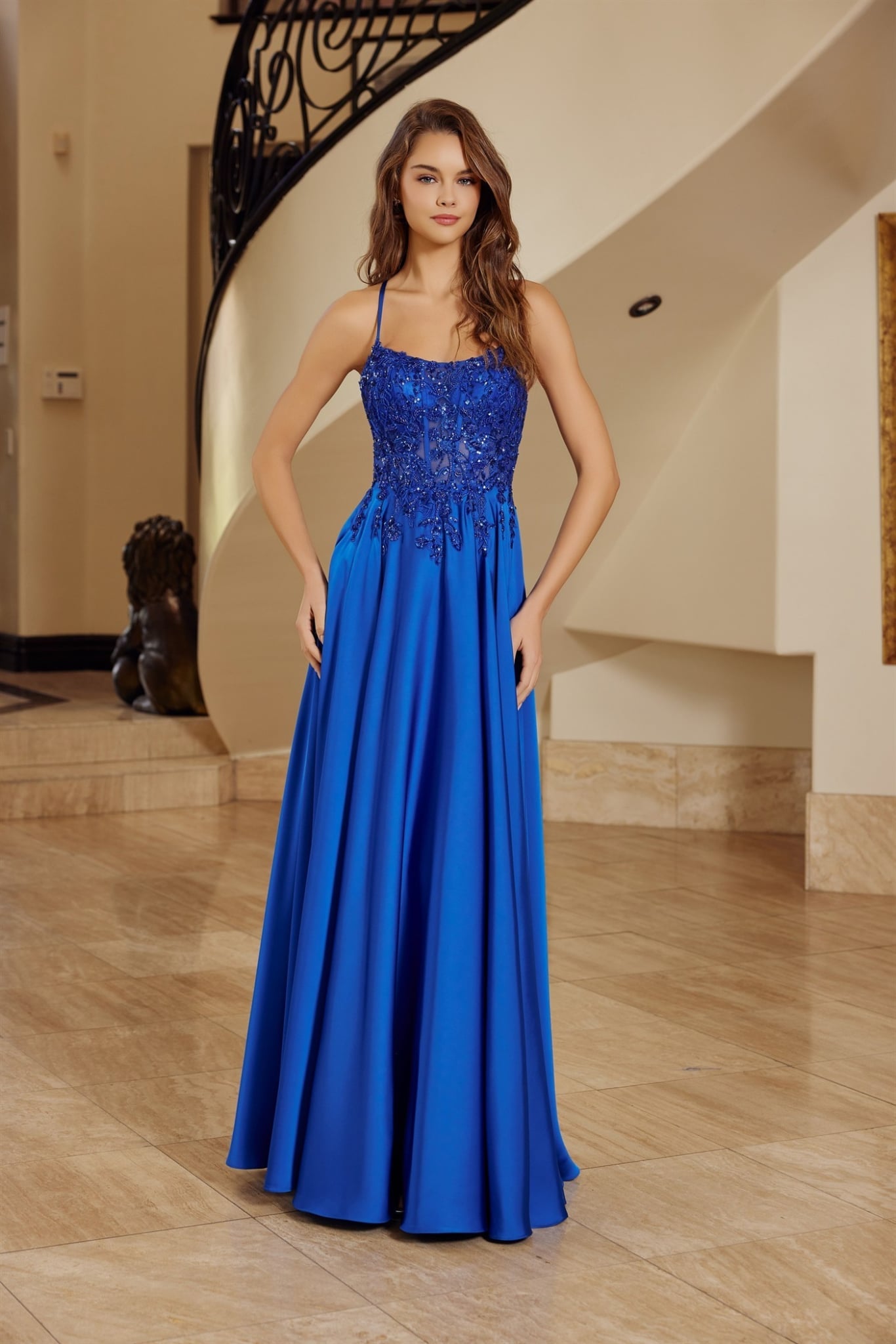 Robe de soirée longue bleu royal, bustier en dentelle florale orné de perles avec fines bretelles et jupe longue fluide en satin, posée devant un escalier luxueux. Modèle disponible en plusieurs couleurs.