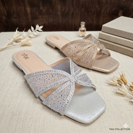 Mules plates de soirée modèle Bella de Taxi Collection. Version argent et version champagne avec brides larges ornées de strass étincelants, bout carré, présentées sur un fond neutre avec fleurs séchées.