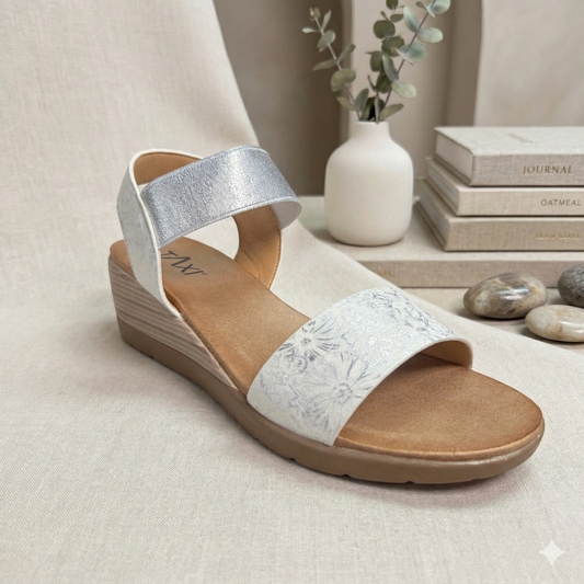 Sandale compensée basse modèle Debbie de Taxi. Bride avant blanche à motifs floraux argentés et large bride élastique argentée à la cheville, présentée sur un fond neutre avec des livres et un vase.