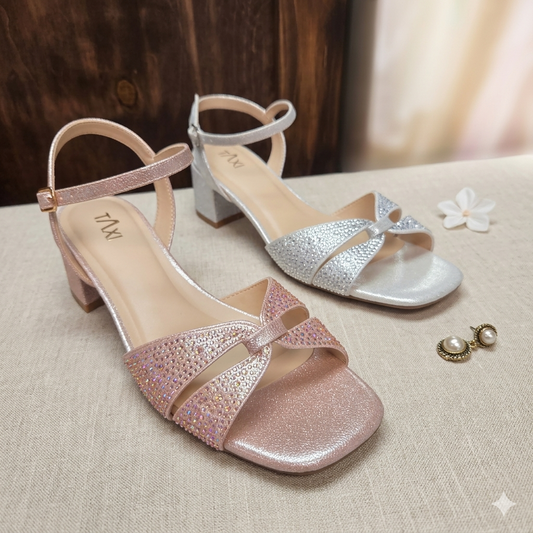 Une paire de sandales Sage de la marque Taxi, une en or rose poudré et une en argent métallique, présentées sur une table en tissu beige. Les sandales ont des brides entrelacées ornées de strass dégradés, des bouts carrés ouverts, et des talons blocs carrés moyens. À côté, une fleur blanche et des boucles d'oreilles en perle. L'étiquette de prix est marquée "TAXICOLLECTION".